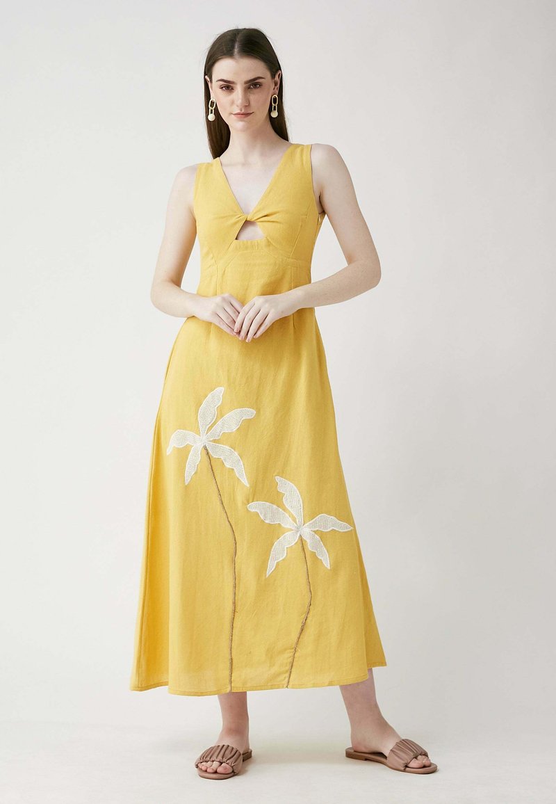 Gelbes Leinen-Maxikleid mit tiefem V-Ausschnitt und Cut-Out-Details. Bestickte weiße Palmblätter zieren den Rock. Tragen mit beigen Sandalen.