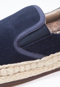 Marinblå mockasinsko utan snörning med elastiska detaljer, beige vävd jute-mellansula och texturerad inre foder. Släta och mjuka ytdetaljer synliga.