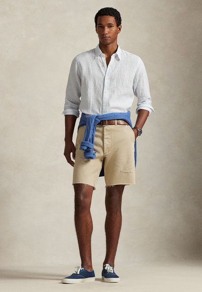 Ανοιχτό μπλε ριγέ πουκάμισο με κουμπιά, μπεζ σορτς με ξεφτισμένες άκρες, μπλε πουλόβερ δεμένο στη μέση και μπλε αθλητικά παπούτσια. Καφέ ζώνη και ρολόι.