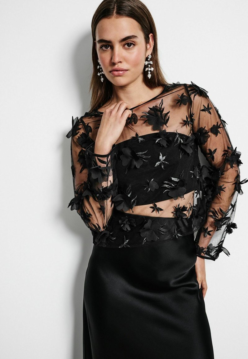 Zwarte cropped top met doorzichtige, geborduurde bloemendetails. Gecombineerd met een elegante, satijnen zwarte rok. Model heeft schouderlang haar.