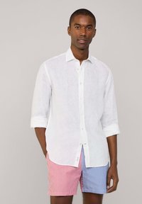 Camicia bianca in lino con maniche arrotolate, abbinata a pantaloni corti a righe rossi e blu. Il design presenta un colletto classico e bottoni frontali.