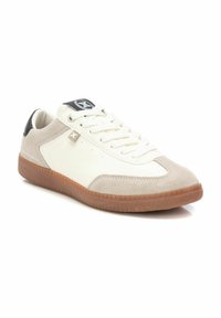Zapatilla blanca con acentos de ante en beige, suela de goma texturizada, diseño de cordones y detalle en negro en el talón. Presenta una marca en la lengüeta.