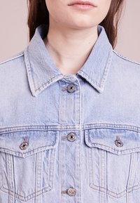 Světle modrá džínová bunda s límcem, stříbrnými knoflíky, dvěma náprsními kapsami a viditelným švem, s klasickým střihem a texturou.