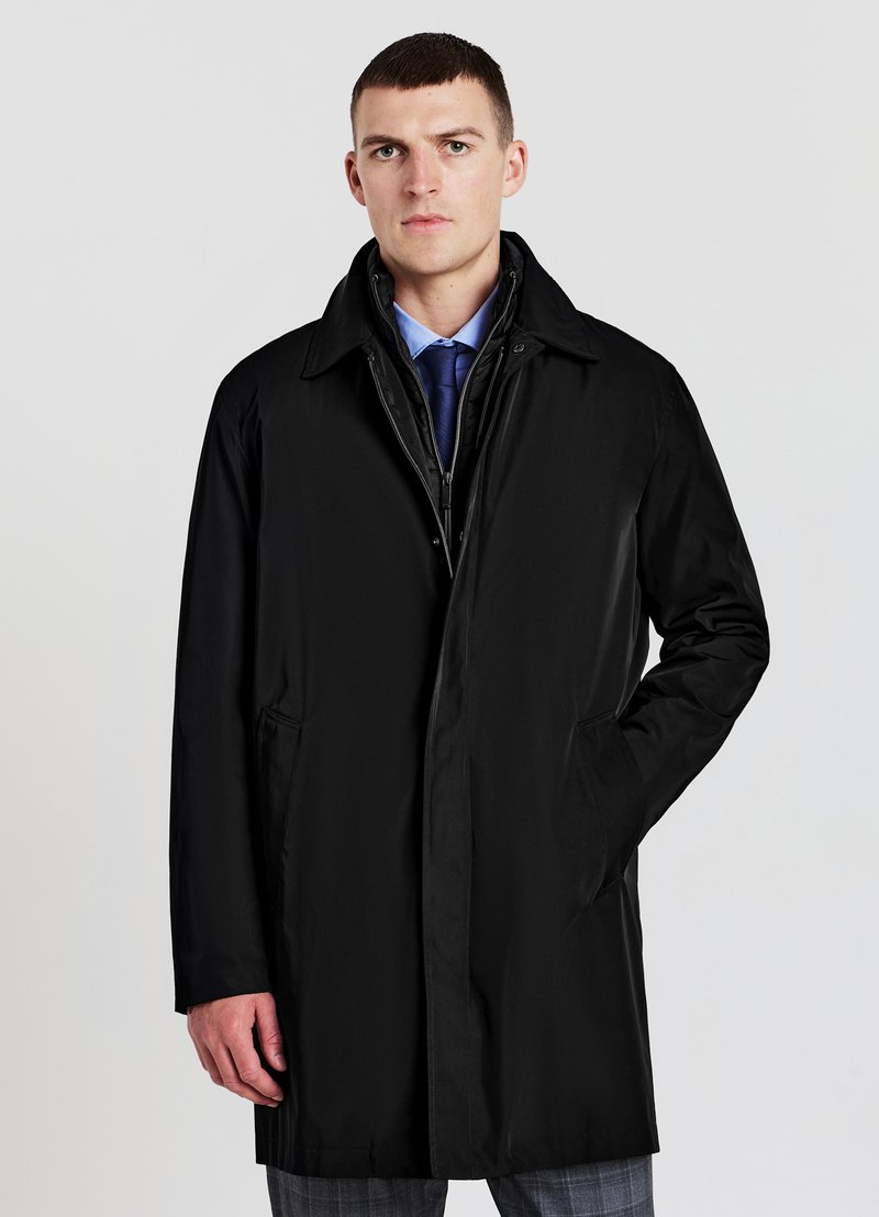 Jeune homme portant un manteau noir, une chemise habillée bleue et une cravate bleu marine, se tenant avec une main dans la poche contre un fond uni.