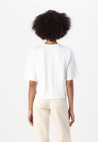 T-shirt bianca cropped con maniche corte e scollatura rotonda, realizzata in cotone, vista di spalle. Abbinata a pantaloni beige chiaro.