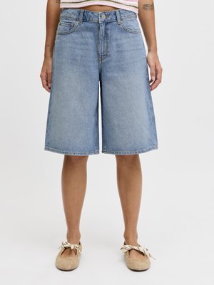 Γυναίκα που φοράει ανοιχτό μπλε τζιν σορτς μέχρι το γόνατο, μπεζ μπαλαρίνες με φιόγκους και κοντό ριγέ τοπ.