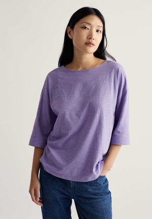 Mladá žena s dlhými čiernymi vlasmi, oblečená v voľnom levanduľovom tričku s tromi štvrťovými rukávmi a modrých džínsoch, s jednou rukou v vrecku.