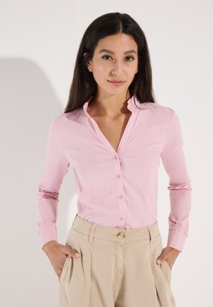 Frau mit langen dunklen Haaren, die ein hellrosa Hemd mit Knöpfen und beige, hoch taillierte Hosen trägt, mit den Händen in den Taschen, stehend vor einem hellen Hintergrund.