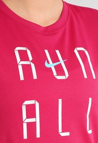 Rosa bomullströja med stora vita bokstäver som stavar "RUN ALL", med en liten blå Nike-logotyp ovanför texten. Rund halsringning.