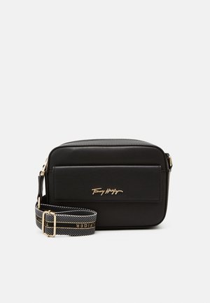 Czarna prostokątna torebka noszona na ramieniu z przednią kieszenią, złotym logo Tommy Hilfiger oraz czarnym regulowanym paskiem z nazwą marki w złotych literach.