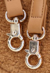 Leren tan riemen met glanzende zilveren metalen clips, met een ronde vorm en gedetailleerde hardware, tegen een gestructureerde bruine stoffen achtergrond.