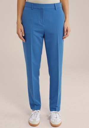 Persoon die blauw op maat gemaakte broek met riemlussen en witte sneakers draagt, staand tegen een neutrale achtergrond.