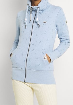 Jasnoniebieska bluza z kapturem zapinana na zamek błyskawiczny, z nadrukami kotwic, kapturem ze sznurkiem oraz kieszeniami na guziki, noszona z jasnymi spodniami.