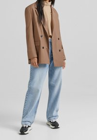 Hnědý dvouřadý blejzr s velkými černými knoflíky, doplněný světle modrými širokými džínami a černobílými teniskami.