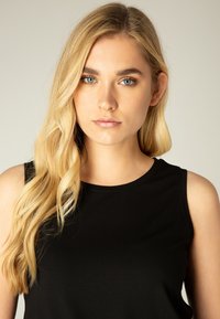 Black sleeveless top, round neckline, fitted design. Long, wavy blonde hair and clear blue eyes are visible against a neutral background.