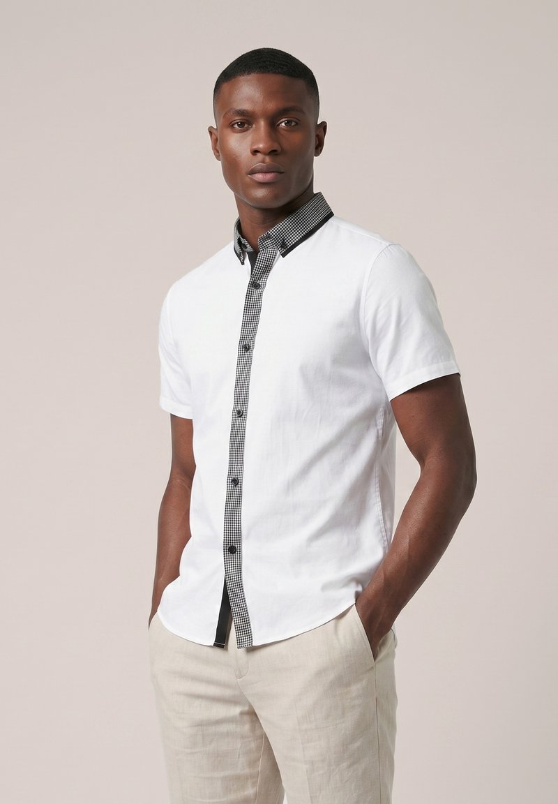 Homme portant une chemise blanche à manches courtes avec une bordure à carreaux noirs et un pantalon beige, debout les mains dans les poches sur un fond uni.