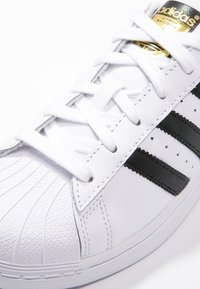 Bílá kožená teniska s texturovanou gumovou špičkou, třemi černými pruhy, zlatým logem a bílými tkaničkami.