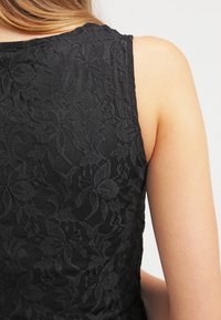 Haut noir sans manches avec motif en dentelle florale porté par une personne à la peau claire et aux cheveux blonds visibles à la nuque.