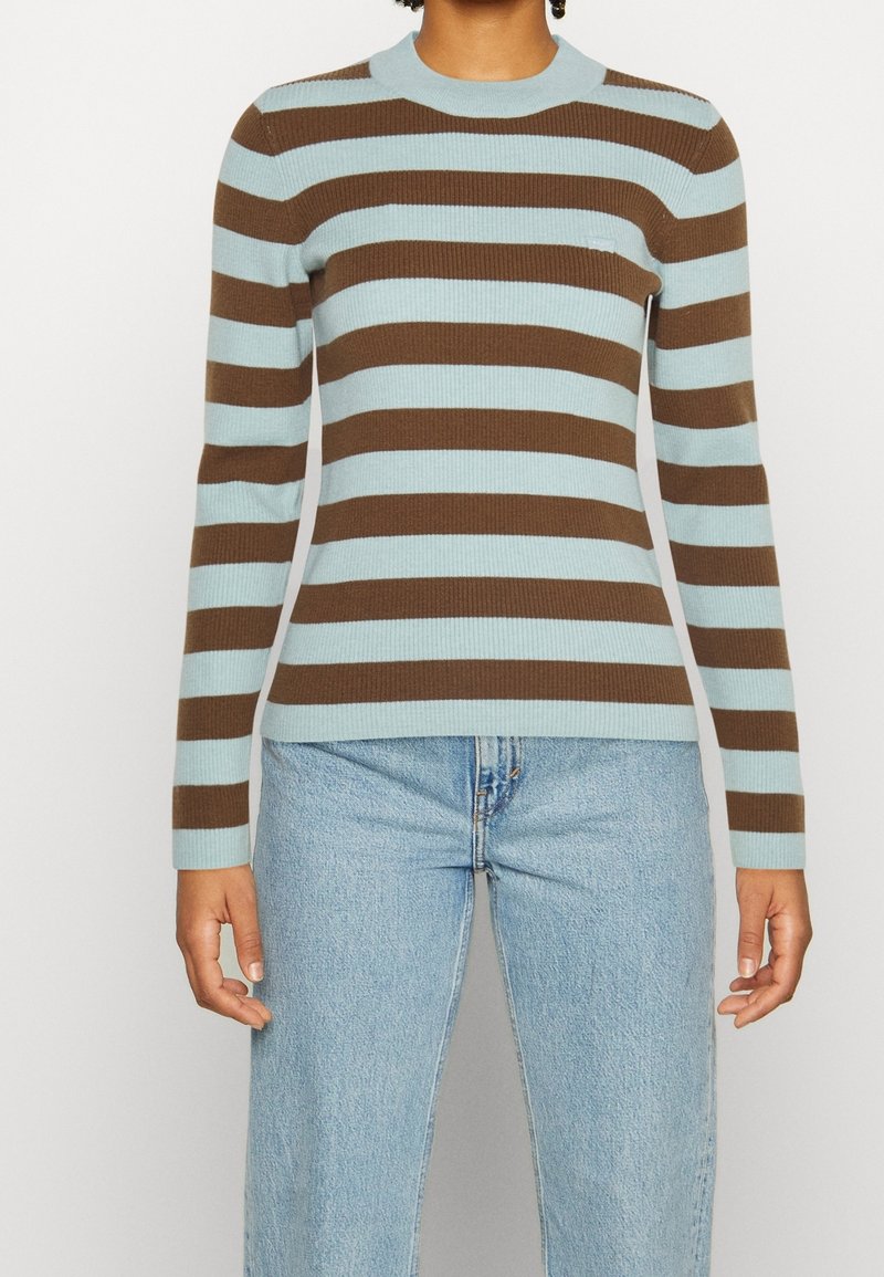 Personne portant un pull en maille ajusté à manches longues avec des rayures horizontales marron et bleu clair, associé à un jean bleu clair.