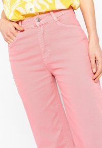 Pantalones de mezclilla rosa con pierna recta, que cuentan con bolsillos frontales, cremallera y cierre de botón metálico. Textura suave con costuras visibles.