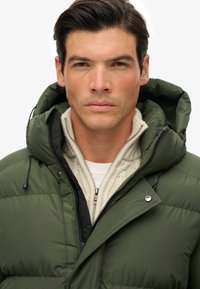 Hombre con cabello oscuro que lleva una chaqueta acolchada verde sobre un suéter de punto trenzado color crema con cremallera, mirando hacia adelante contra un fondo blanco.