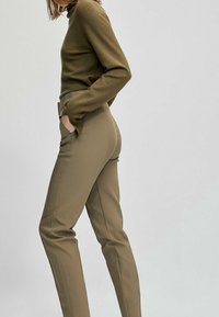 Un pull à col roulé en tricot vert olive et un pantalon beige ajusté avec poches latérales et coupe slim, ornés de coutures discrètes le long des jambes.