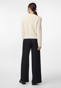 Cremfarbener Pullover mit gewelltem Saum, kombiniert mit schwarzen weit geschnittenen Hosen. Der Pullover hat ein erhabenes Muster und lange Ärmel.
