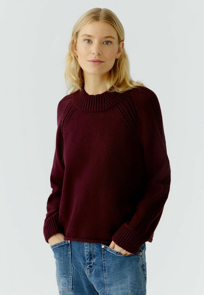 Burgundy knit sweater with a ribbed crew neck, textured shoulder seams, and wide sleeves, paired with light blue denim jeans.