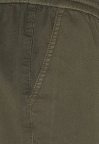 Olivově zelená tkanina s hladkým povrchem, s elastickým pasem a boční kapsou. Šité detaily podél švů.