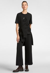 Camiseta negra con un diseño de lentejuelas, combinada con pantalones negros holgados. El conjunto incluye un suéter negro atado para un toque adicional de capas.