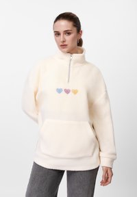 Sudadera de forro polar color crema con cuello alto y media cremallera. Presenta tres corazones bordados en colores pastel y un bolsillo delantero.