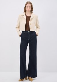 Beige kort blazer med strukturerad tyg och tre knappar, matchad med mörkblå högmidjade vida jeans med kontrastsömmar och fickor.