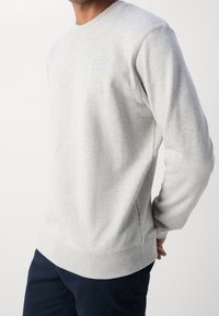 Szara bluzka z bawełnianego materiału, okrągły dekolt, ściągacze przy mankietach i na dolnym brzegu. Posiada małe, kwadratowe logo na piersi. Dopasowany krój.