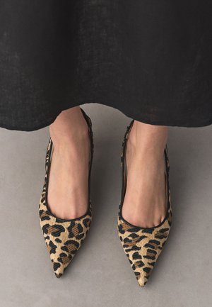 Füße, die spitz zulaufende flache Schuhe mit Leopardenmuster tragen, mit einem schwarzen Rock, der über den Knöcheln sichtbar ist, vor einem grauen Fußboden-Hintergrund.