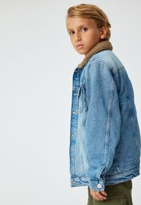 Ljusblå denimjacka med en krage i brun fuskpäls, knappstängning fram och sidofickor. Designen har en avslappnad passform och synlig sömnad.