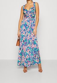 Maxi mekko eläväisessä sinisessä kukkakuviolla, jossa on tiukka vartalo-osa, leveät hihnat ja virtaava hame. Ruskea käsilaukku ja remmikenkiä.