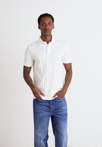 Hvit polo skjorte med teksturert mønster, korte ermer og liten krage, i kombinasjon med lys blå denimjeans. Modellen står med hendene i lommene.