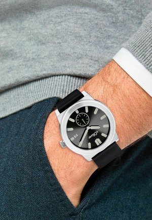 Armbanduhr mit schwarzem Ziffernblatt, silbernem Gehäuse und schwarzem Lederarmband, das am linken Handgelenk getragen wird, kombiniert mit einem grauen Pullover und einer dunkelblauen Hose.