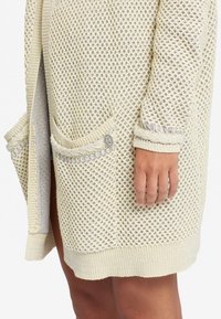 Stickad cardigan i ljus grädde med ett nätmönster, som har två framfickor och subtila grå accenter vid fickkanterna och ärmsluten.