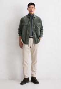 Camicetta in velluto a coste verde con due tasche sul petto, indossata sopra un maglione aderente scuro a collo alto. Pantaloni in velluto a coste beige chiaro e stivali alla caviglia in camoscio scuro.