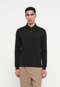Polo shirt nera a maniche lunghe con colletto a coste e patta con tre bottoni, e piccolo logo a sinistra sul petto. Tessuto liscio.