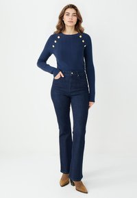 Pull en maille bleu marine avec boutons dorés sur les épaules, associé à un jean flare en denim foncé taille haute et des bottines à talons carrés couleur camel.