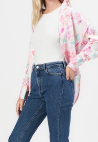 Kvinna som bär blå högmidjade jeans, vit ribbad tanktop och en öppen pastellrosa blommig skjorta med en hand i fickan.