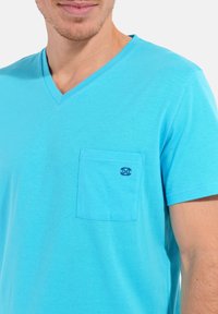 T-shirt à col en V, à manches courtes, de couleur turquoise vif avec une poche avant arborant un petit logo bleu foncé. Tissu doux en coton.