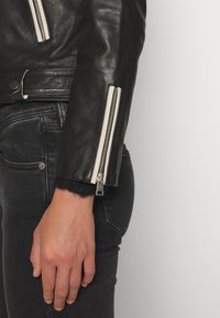 Chaqueta de cuero negra con cremalleras plateadas, acabado texturizado y un detalle de puño de lana gris en la muñeca. Jeans de mezclilla negros en el fondo.