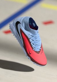 Nike athletic shoe with a red and black textured upper, light blue laces, and a patterned sole. Features a rounded toe and a low profile.