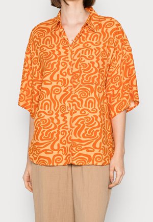Vrouw draagt een losvallend, kort mouwig overhemd met oranje abstracte swirl-patronen en beige broek tegen een effen achtergrond.