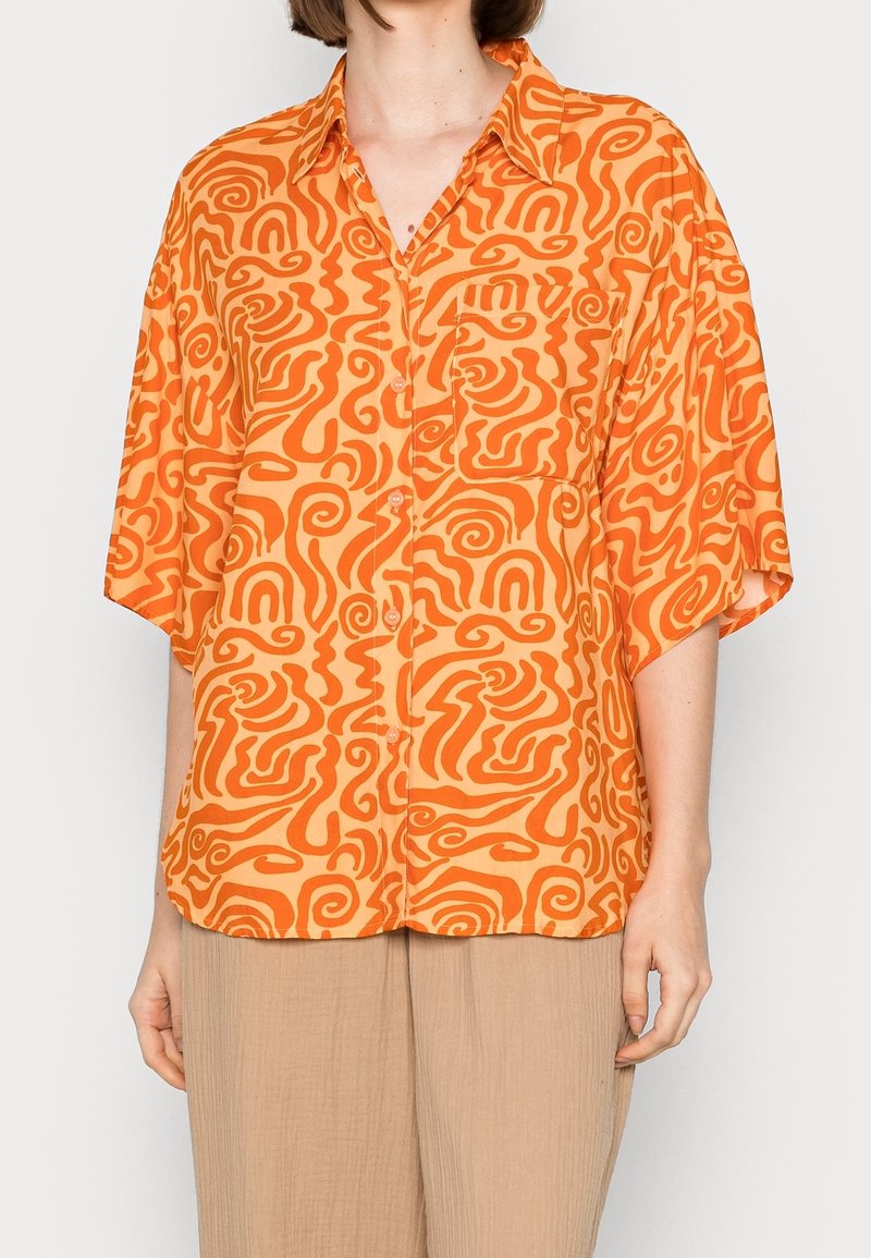 Une femme porte une chemise ample à manches courtes avec des motifs abstraits en spirale orange et un pantalon beige, sur un fond uni.
