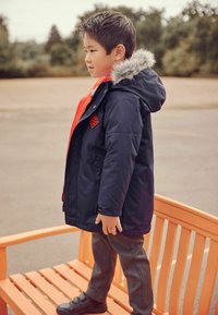 Granatowy płaszcz zimowy z kapturem wykończonym futrem, czerwonymi akcentami, z zamkiem błyskawicznym i emblematem, w połączeniu z szarymi spodniami i czarnymi butami.