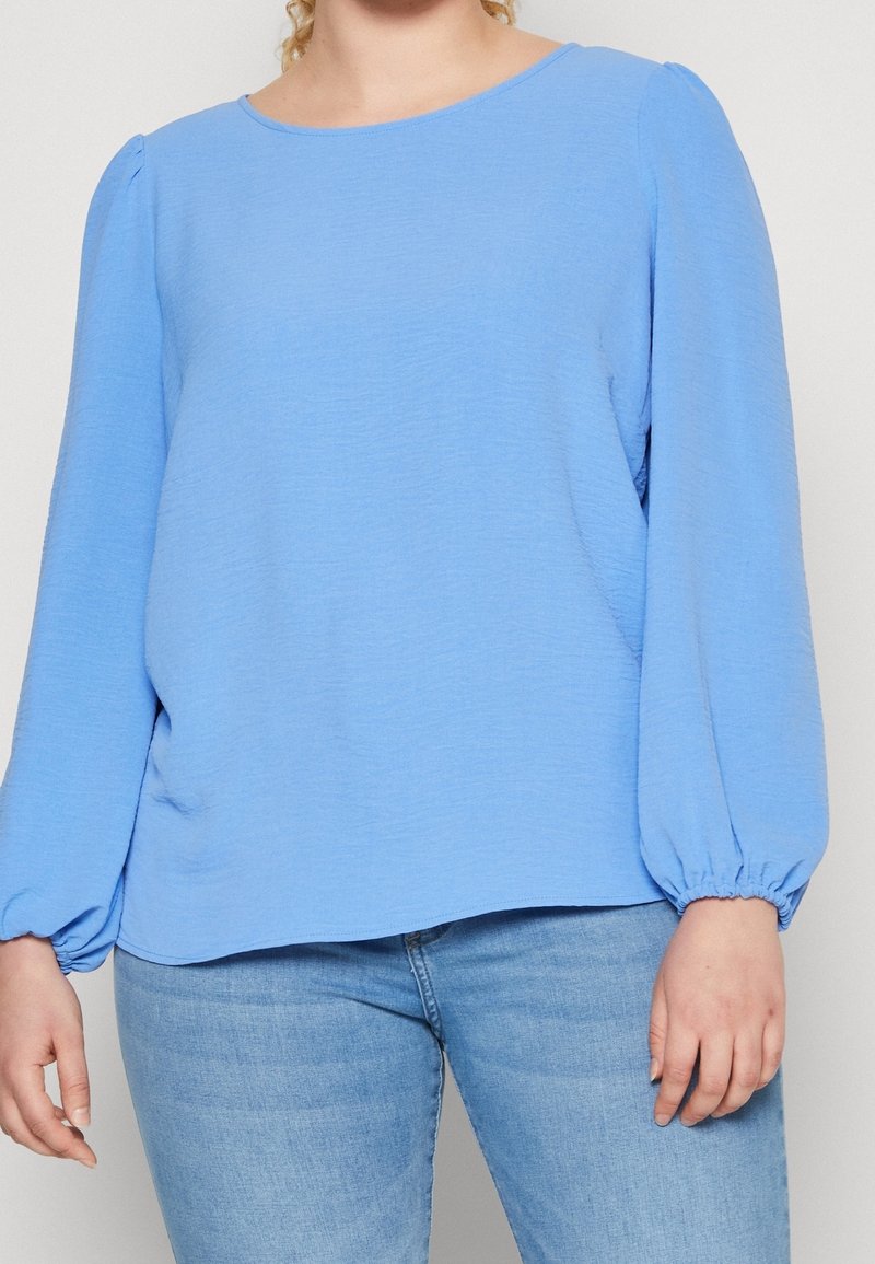 Top azzurro chiaro a maniche lunghe con scollatura rotonda e polsini arricciati, realizzato in tessuto testurizzato; vestibilità comoda con pieghe laterali.
